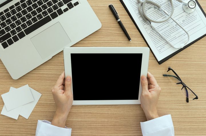 Doctor holding a tablet with a blank screen, next to a laptop, stethoscope, and glasses.