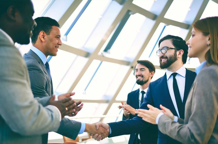 Group of businessmen shaking hands in an office, smiling, while others applaud.