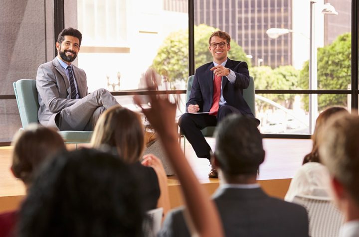 Two businessmen on stage while the audience raises their hands.