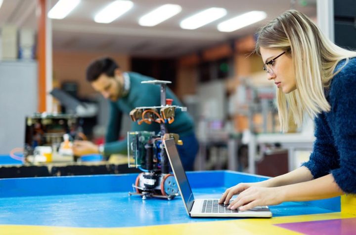 Woman working on a laptop with a small robot in front of her.