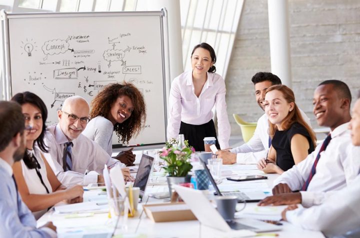 Team of people in an office discussing a project, smiling woman standing by a whiteboard.