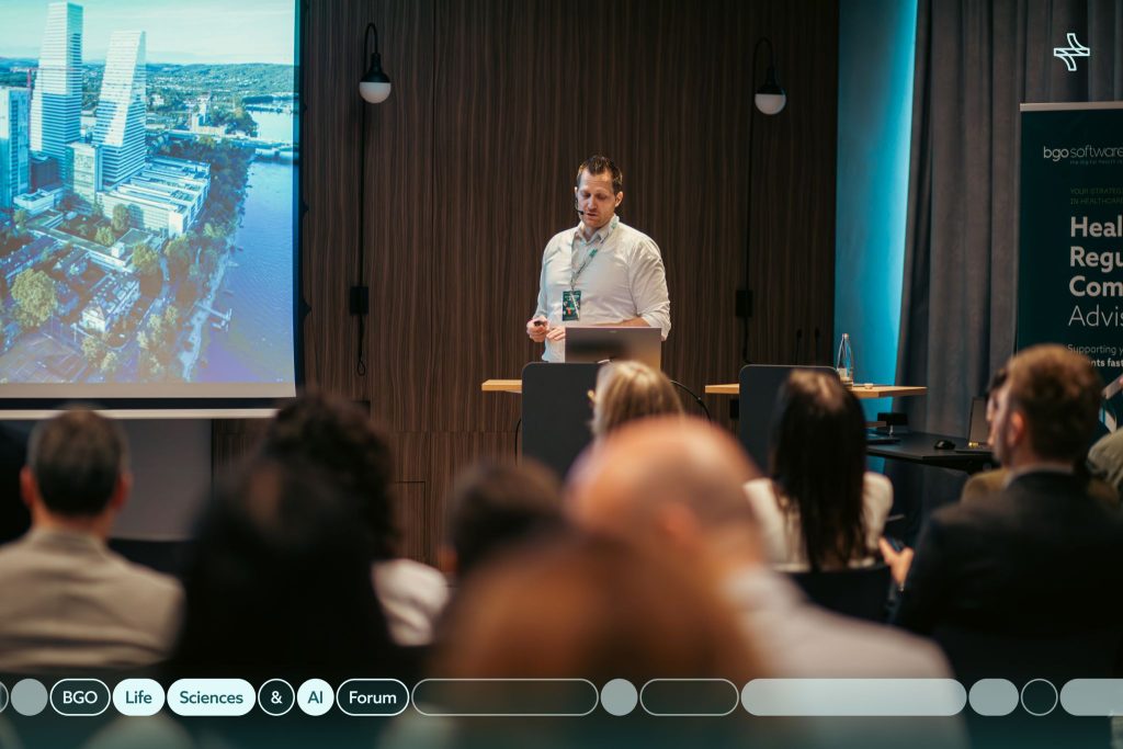 A speaker presenting on stage at the BGO Life Sciences and AI Forum, talking about healthcare regulatory compliance to an engaged audience in a conference hall.